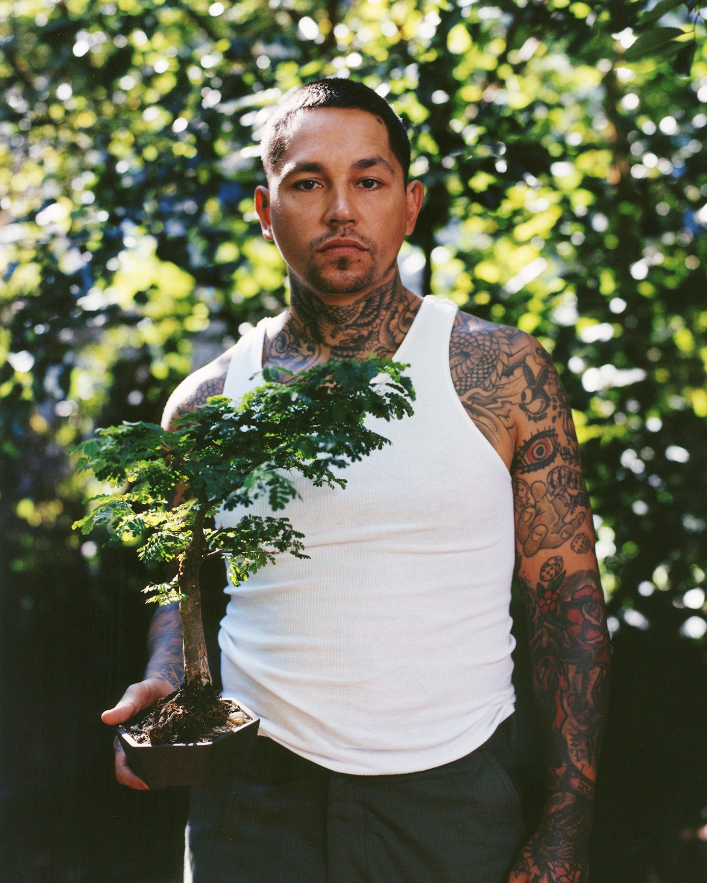 tattooed man in tank top holding bonsai tree outside